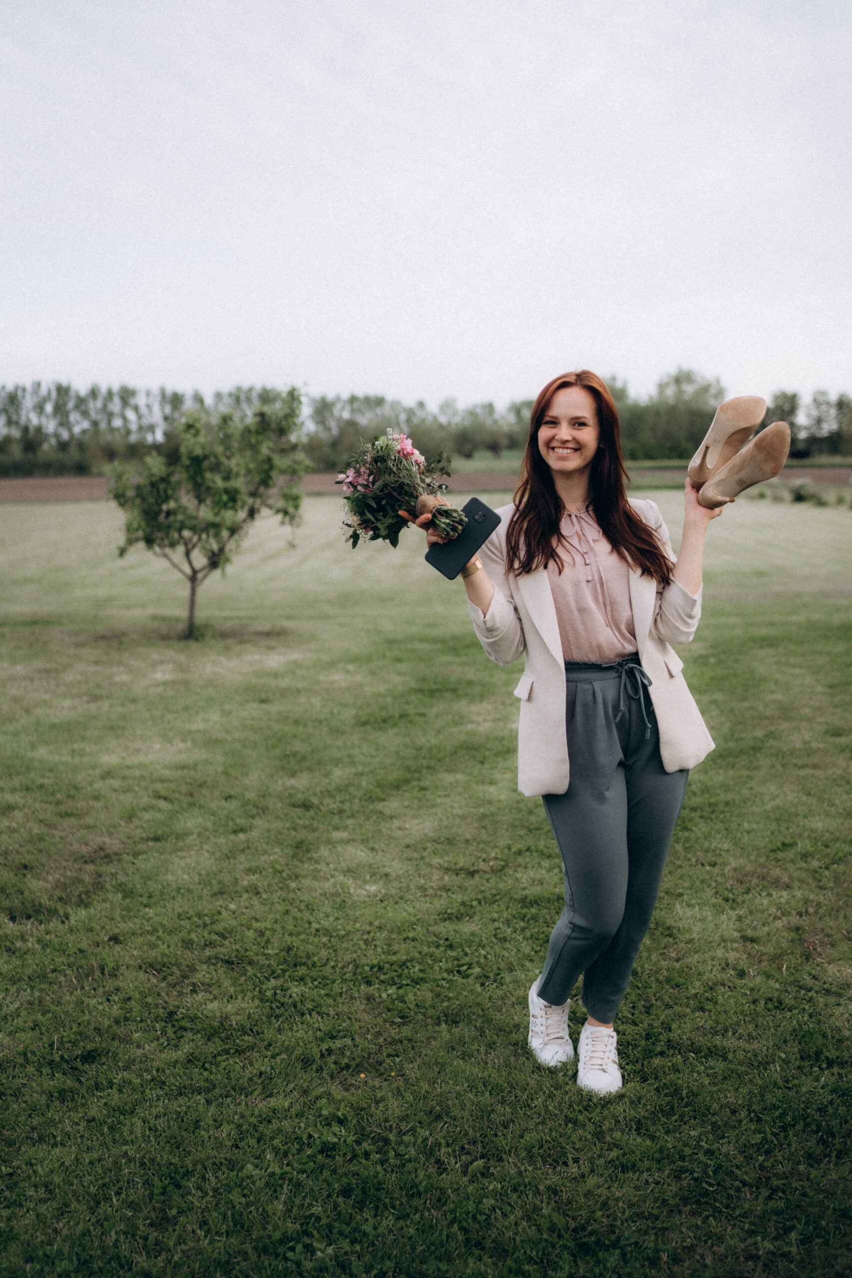Hochzeitsplanerin Romina Blume hält beim Paarshooting im Garten die Schuhe und den Brautstrauß der Braut und lacht fröhlich