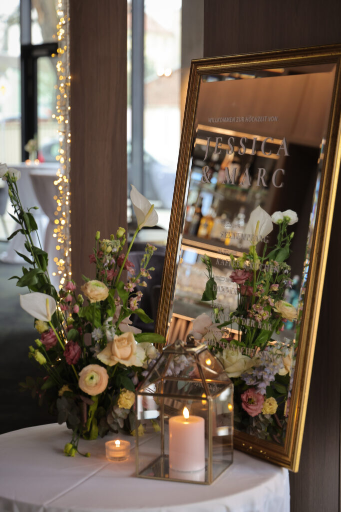 Willkommensschild aus Spiegel mit Goldrahmen, dekoriert mit Kerzen und floralen Gestecken im Vintage-Stil.