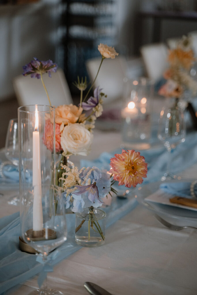 Detail einer sommerlichen Hochzeitstafel mit pastellfarbenen Blumen, Kerzen und hellblauer Tischdekoration.